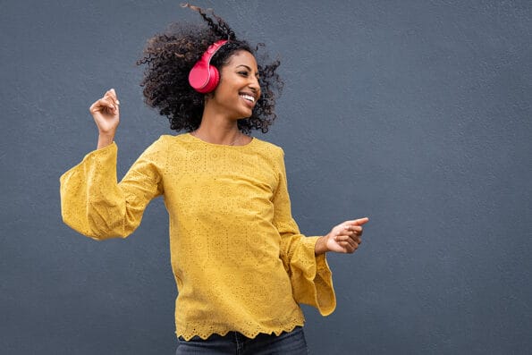 Beautiful black girl listening music. Cheerful young woman with curly hair making dance moves to the rhythm of music while listening through wireless headphones isolated on grey wall.