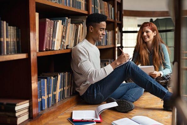 Smiling classmates doing homework in a college library. University students preparing an essay or exam together. Happy young woman and african guy sitting on floor in school library have discussion.