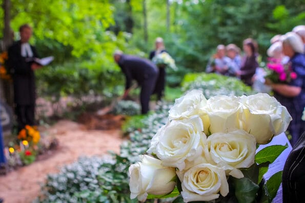 Roses in cemetery with people in the background. Funeral in cemetery; White roses in cemetery. Prayer at the tomb. Funeral ceremony. White roses at funeral near the grave.