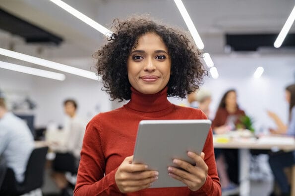 Portrait of happy multiethnic businesswoman using digital tablet in agency. Successful business woman in casual clothing working on tablet. Mixed race young woman looking at camera in creative office.