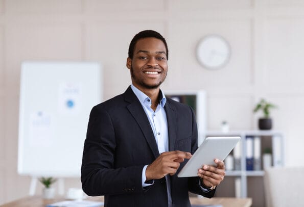 Smiling black entrepreneur young man holding digital tablet, using modern technologies in business, office interior, copy space. Cheerful african american businessman using app for business
