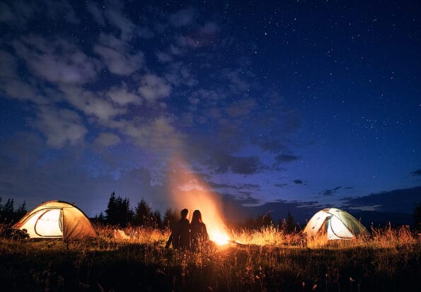 camping lantern Fantastic view of night starry sky over grassy hill with illuminated camp tents, campfire and hikers. Young tourists having a rest near bonfire. Concept of hiking, night camping and relationship.