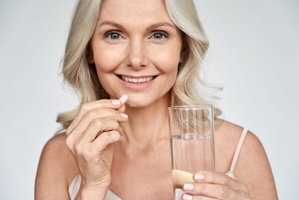 Smiling happy healthy middle aged 50s woman holding glass of water taking dietary supplement vitamin pink pill isolated on white background. Old women multivitamins antioxidants for anti age beauty.