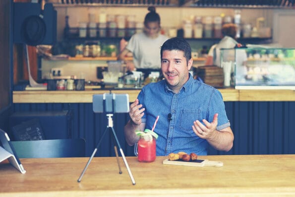 Social media influencer or food blogger creating content inside small restaurant - man sharing online food review using smartphone on tripod and lavalier, smiling content creator vlogger filming video
