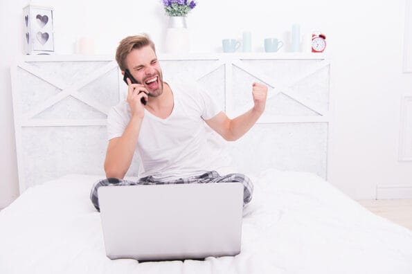 Its a deal. Happy man make winner gesture in bed. Mobile communication. Phone conversation. Computer technology. Bet winner. Online betting. Virtual casino games.