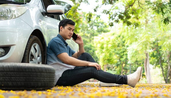 Asian man sitting beside car and using mobile phone calling for assistance after a car breakdown on street. Concept of vehicle engine problem or accident and emergency help from Professional mechanic
