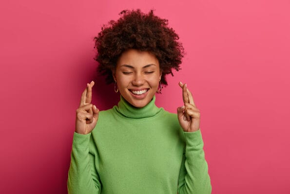 luck Hopeful happy woman smiles positively, makes wish and confident to win, crosses fingers for good luck, eagerly wishing success, closes eyes, wears green turtleneck, isolated over vibrant pink wall