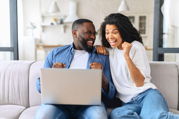 games, nft token, Overjoyed cheerful multiracial couple celebrating good news looking at the laptop screen, young excited woman and man scream happily sitting with computer on the cozy couch at home, won in video game