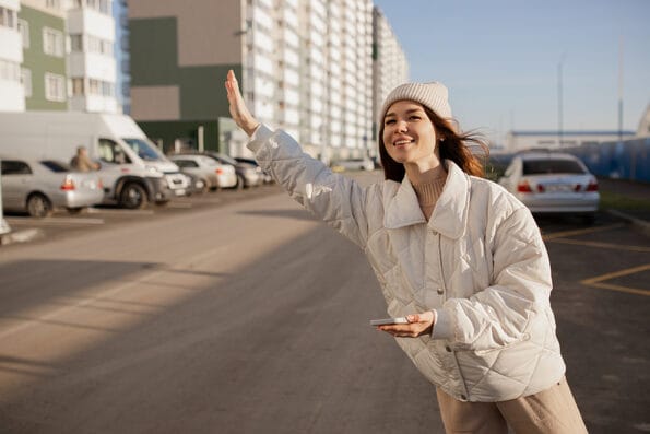 A young girl in a white jacket waves a taxi, which she called through the app. A young girl catches a taxi with a phone in her hands. A joyful girl catches a taxi at a bus stop.