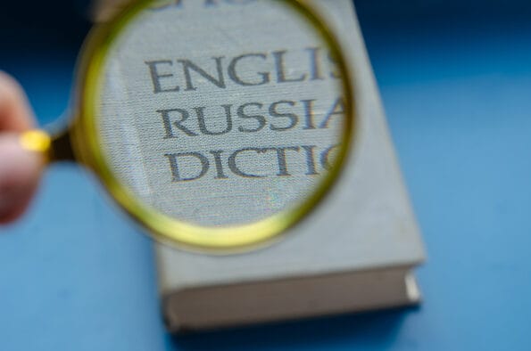 Education, foreign language learning, search for knowledge concept. Magnifying glass and hardcover book on blue background. English-Russian language dictionary enlarged in the loupe.