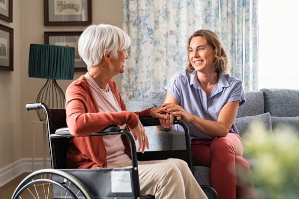 Mature woman comforting senior mom sitting on wheelchair at nursing home. Cheerful woman talking to old disabled mother at elder care centre. Loving caregiver taking care of elderly woman at home.