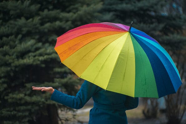 A girl in a green coat hides under an umbrella of rainbow colors from the rain. The girl removed her hand from under the umbrella to learn about the end of the rain.