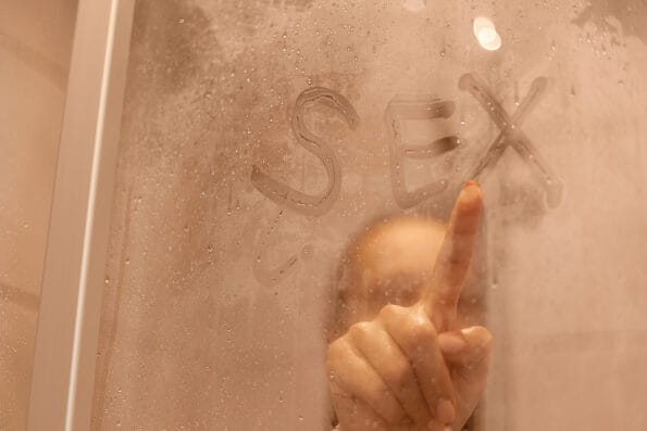 Portrait of beautiful woman taking a shower and writing word sex on the sweat and blurred glass, attractive female doing morning hygiene procedures, body care.