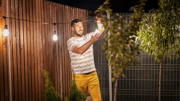 Party time in backyard with happy millennial man hanging string lights in trees - Weekend night mood with smiling guy arranging the light garland for outdoor dinner party in home garden