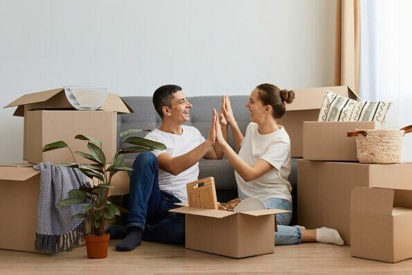 Family moved in new home, sitting on the cough surrounded with cardboard boxes, holding hands, giving five, rejoicing to move in together, relocated in new flat.