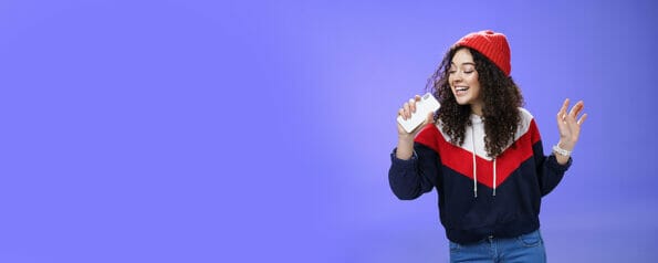 Attractive happy young woman with curly hair in hat singing enjoying perfect winter day singing along in smartphone, holding mobile phone like microphone, adore karaoke over blue background